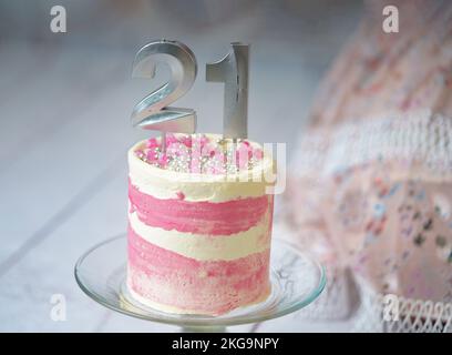 21. Geburtstagskuchen, rosa und silberne Kuchen mit ein paar Streuseln und 21. Kerzenlicht mit einem Frauenkleid im Hintergrund. Stockfoto