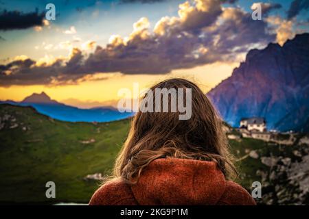 Beschreibung: Sportliche Frau in Outdoor-Jacke sieht sich die wunderschöne Bergkulisse bei Sonnenuntergang am Rifugio Passo Valparola an. Falzarego-Pass, Dolomiten, Süd-Tir Stockfoto