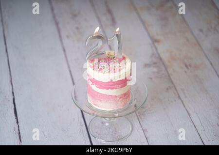 21. Geburtstagskuchen, rosa und silberne Torte mit ein paar Streuseln und 21. Kerzenlicht auf weißem Holzhintergrund. Stockfoto