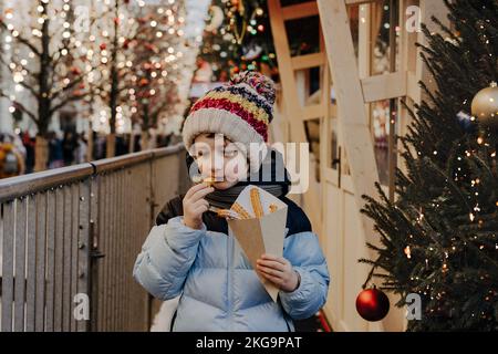 Niedlicher kleiner Junge, der auf dem traditionellen Weihnachtsmarkt im Schnee Churros isst. Das Kind genießt Süßigkeiten auf dem Weihnachtsmarkt. Winterurlaub mit der Familie. Frohe Stockfoto