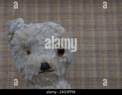 Klassischer Teddybär in liebenswertem, getragenem Zustand mit fehlendem Auge, fehlendem Ohr und fehlendem Fell. Nahaufnahme des Gesichts mit kariertem Kofferdeckel. Stockfoto