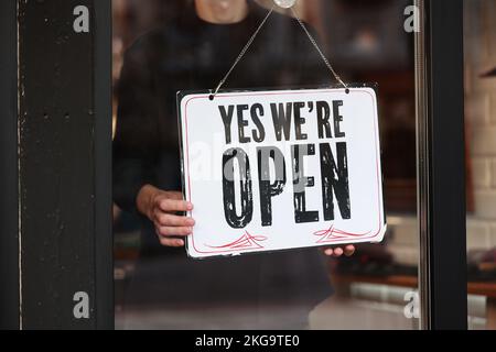 Der Besitzer hängt eine Tafel mit Inschrift auf. Ja, wir haben geöffnet bei Glastürladen, Café, Schönheitssalon, Friseurladen nach Coronavirus-Quarantäne. Das Geschäft wird wieder eröffnet. Stockfoto