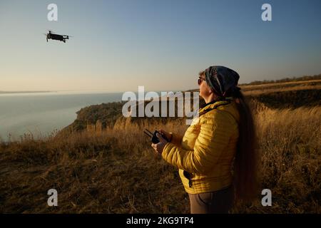 Hübsche Frau, die Drohne mit Herbstgras bei Sonnenuntergang steuert. Stockfoto