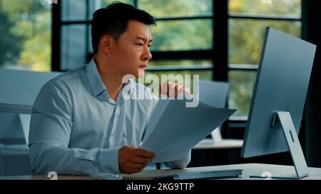 Verzweifelter, trauriger, asiatischer, japanischer Geschäftsmann, der im Büro sitzt, liest Dokumente, fälscht die Benachrichtigung bei der Recherche von Geschäftsdokumenten Stockfoto
