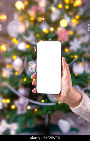 Nahaufnahme des Smartphones mit leerem Bildschirm-Mockup im Hintergrund des Weihnachtsbaums, Kunde hält Telefon in der Hand, online einkaufen, GI kaufen Stockfoto