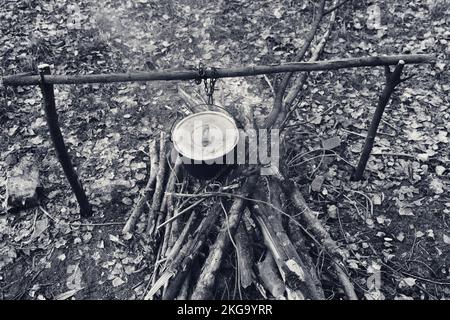 Kochen in altem Ruß-Kessel am Lagerfeuer der Lichtung. Weitwinkelblick von oben. Schwarzweiß-Retro-Bild. Stockfoto