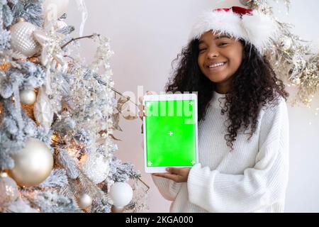 Lächelndes schwarzes Mädchen in Weihnachtsmann Hut zeigt Tablette mit grünem Bildschirm, hält Chroma-Key-Gadget in der Nähe von Weihnachtsbaum, Werbung Weihnachtsartikel, Apps Stockfoto