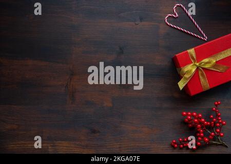 Rote Geschenkbox mit goldenem Band auf dunklem Holzhintergrund mit Zweigen aus roten Beeren und einem Herz aus Zuckerrohr, weihnachtlicher Hintergrund Stockfoto