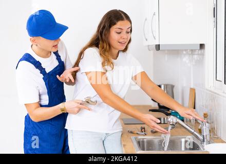 Glückliche, zufriedene Hausfrau überprüft den Betrieb des Wasserhahns in der Küche und danke dem Klempner Stockfoto