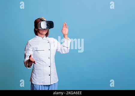 Positiver leitender Koch trägt eine Virtual-Reality-Schutzbrille während der Zubereitung kulinarischer Mahlzeit arbeitet an einem gesunden Rezept. Freundlicher Koch mit Restaurant, einheitlicher Koch, Gastronomie, Speisengericht. Lebensmittelindustrie Stockfoto