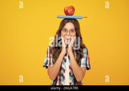 Zurück zur Schule. Teenager Schülerin mit Apfel auf dem Kopf, bereit zu lernen. Schulkinder auf isoliertem gelben Hintergrund. Schockiert überrascht Teenager Mädchen Stockfoto