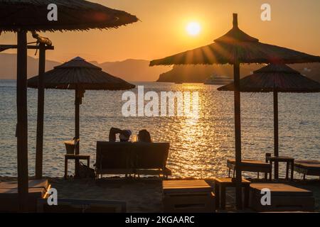 IOS, Griechenland - 13. September 2022 : Blick auf ein Paar, das auf Sonnenliegen liegt, Wein trinkt und den herrlichen Sonnenuntergang in Orange in iOS Greece genießt Stockfoto