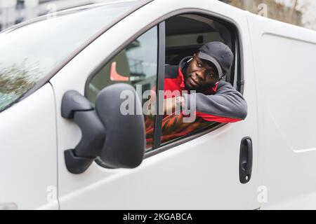 Ein hübscher bärtiger schwarzer Liefermann mit schwarzer Baseballmütze und roter Jacke, der aus dem Fenster seines weißen Lieferwagens guckt. Paketzustellkonzept. Hochwertiges Foto Stockfoto