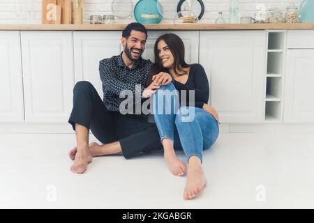 Ein junges, fröhliches, stilvolles Paar, das lacht, sitzt zusammen auf dem weißen Küchenboden in ihrem neuen hellen Apartment. Hochwertiges Foto Stockfoto