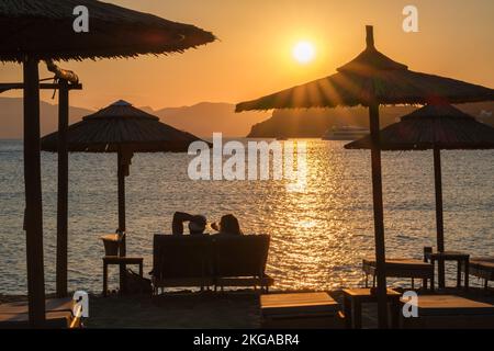 IOS, Griechenland - 13. September 2022 : Blick auf ein Paar, das auf Sonnenliegen liegt, Wein trinkt und den herrlichen Sonnenuntergang in Orange in iOS Greece genießt Stockfoto