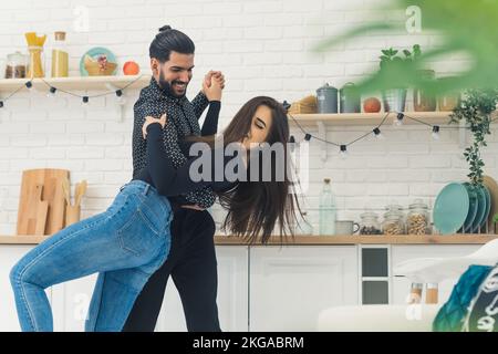 Wunderschöne junge Frau mit langen braunen Haaren und einem fröhlichen, eleganten Mann, der zusammen lächelt und tanzt, Händchen hält, in der Küche in ihrer neuen hellen Wohnung, um den Umzug zu feiern. Hochwertiges Foto Stockfoto