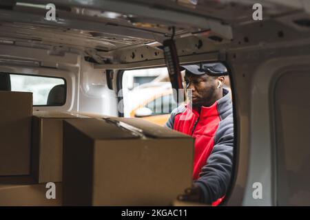 Fokussierter afroamerikanischer Arbeiter - Bote - holt wertvolle Papppakete für seine Kunden. Lieferkonzept für Online-Shopping. Van POV. Hochwertiges Foto Stockfoto