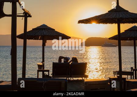 IOS, Griechenland - 13. September 2022 : Blick auf ein Paar, das auf Sonnenliegen liegt, Wein trinkt und den herrlichen Sonnenuntergang in Orange in iOS Greece genießt Stockfoto