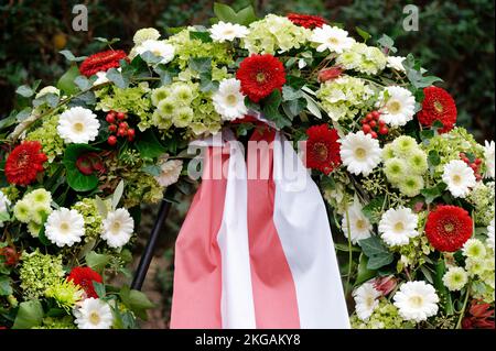 Bestattungskranz auf einem Friedhof mit bunten Blumen und rot-weißer Schleife auf einem Metallrahmen Stockfoto