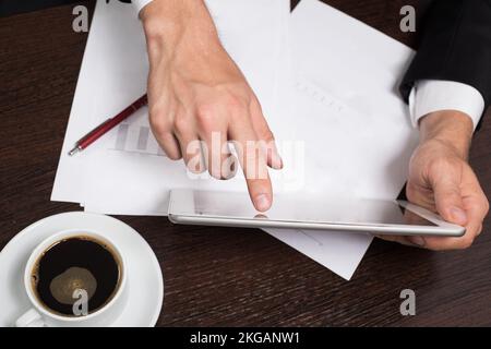 Geschäftsmann im Business-Anzug verwendet ein Tablet. Auf dem Tisch Becher Café, Mobil, Stift Stockfoto