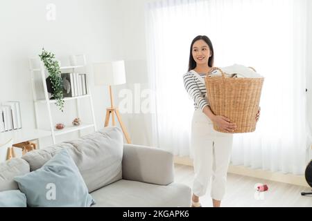 Eine junge Asiatin, die gerne dreckige Klamotten in den Korb im Wohnzimmer mitnimmt. Schöne Reinigungskräfte, die sauber gemacht und geputzt haben. Hausarbeit- und Hausarbeit-Konzept. Stockfoto