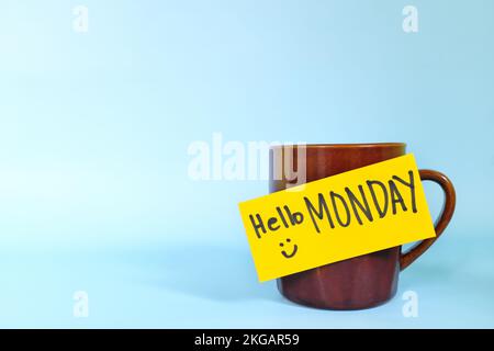 Willkommen, hallo und Happy Monday Concept. Selektiver Fokus der Kaffeetasse mit hellgelbem Papierzettel und schriftlicher Nachricht isoliert auf blauem Hintergrund. Stockfoto