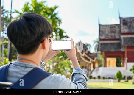 Attraktiver junger asiatischer Reisender oder Rucksacktourist, der mit seinem Smartphone ein Foto des wunderschönen nordthailändischen Tempels macht. Modell des weißen Bildschirms des Telefons. H Stockfoto