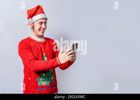 Ein glücklicher Kerl mit weihnachtsmütze, der isoliert am Handy lächelt. Menschen mit weihnachtsmann-Hut, die ein Handy benutzen und lächeln, isolierter, lächelnder junger Mann zu weihnachten Stockfoto