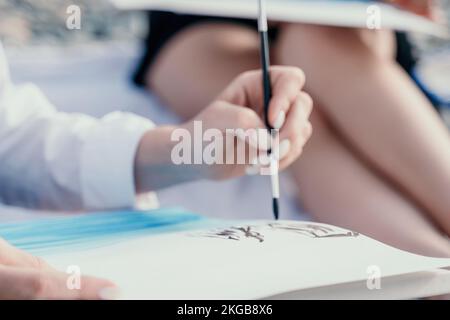 Unverkennbare Hände einer Frau, die ihr eigenes Aquarell mit Meeresstrand hält. Amateur-Maler. Kreative Künstlerin zeichnet das Bild am Stockfoto