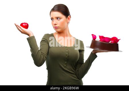 Zwingen Sie mich nicht zu wählen. Studiofoto einer jungen Frau, die sich zwischen einem Apfel und einem Kuchen entscheidet, isoliert auf Weiß. Stockfoto