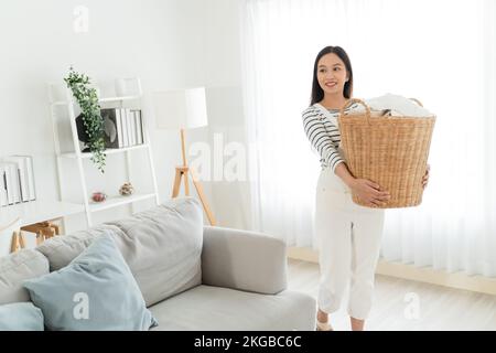 Eine junge Asiatin, die gerne dreckige Klamotten in den Korb im Wohnzimmer mitnimmt. Schöne Reinigungskräfte, die sauber gemacht und geputzt haben. Hausarbeit- und Hausarbeit-Konzept. Stockfoto