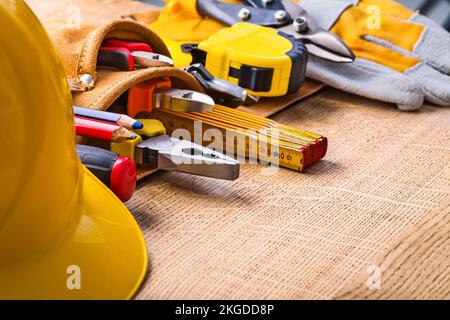 Großer Satz von Bauwerkzeugen in Werkzeuggürtel Harthelm-Schraubendreher Bleistiftspitzer Zange Bandmaß Cutter Handschuhe auf Holzplatte Stockfoto