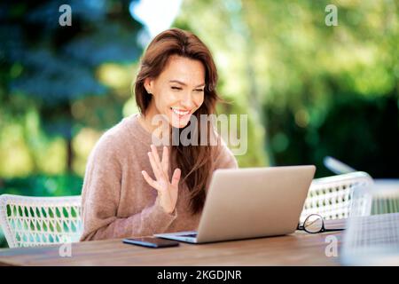 Selbstbewusste Frau mittleren Alters mit Laptop, die auf dem Balkon sitzt und ein Notebook benutzt. Attraktive Frau, die während eines Videogesprächs mit der Hand winkt. Stockfoto
