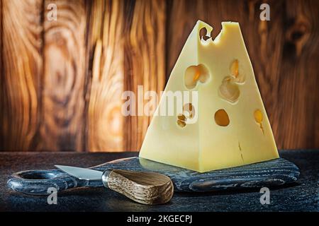 maasdamer Käse, großes Stück und Messer auf kleinen gebogenen Brettern, alter Holzhintergrund Stockfoto