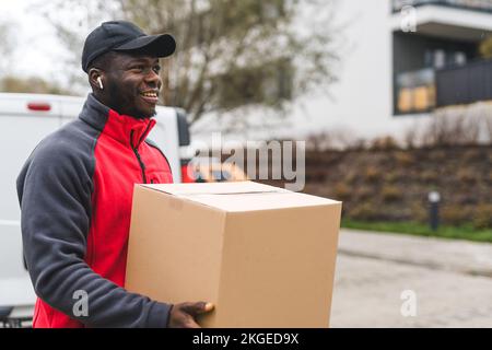 Schwarzer junger Erwachsener trägt Arbeitskleidung, roter, lächelnder Pullover und trägt Pappkartons zur Lieferung. Horizontalschuss im Freien. Hochwertiges Foto Stockfoto