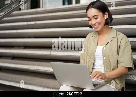 Remote-Mitarbeiter. Ein lächelndes asiatisches Mädchen sitzt draußen auf der Straße mit einem Laptop. Glückliche junge Frau, die aus der Ferne am Computer arbeitet, Musik kabellos hören Stockfoto