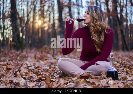 Wunderschöne junge indische Frau in Übergröße, die bei Sonnenuntergang ein Glas Rotwein im Eichenwald genießt Stockfoto