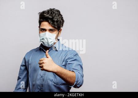 Portrait eines selbstbewussten Mannes in einer medizinischen Maske in grauem Hemd, der Daumen-hoch zeigt und die Kaemra ansieht. Porträt eines Mannes mit OK-Zeichen. Stockfoto