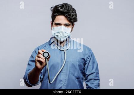 Lass mich nachsehen. Selektiver Fokus auf das Diaphragma eines Stethoskops in der Hand eines männlichen Arztes, der eine Maske auf grauem Hintergrund trägt. Stockfoto