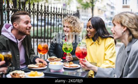 Multirassische glückliche junge Freunde essen und trinken, verschiedene fröhliche Freunde lachen beim Essen, Spaß zusammen am Restauranttisch Stockfoto