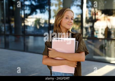 Lächelndes Mädchen, das auf der Straße steht und Notizbücher hält. Kinder im Freien. Zurück zum Schulkonzept Stockfoto