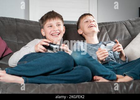 Portrait von zwei erstaunlichen glücklichen barfuß jungen Kindern, die zu Hause auf einem grauen Sofa sitzen, den Controller-Joystick halten und Videospiele spielen Stockfoto