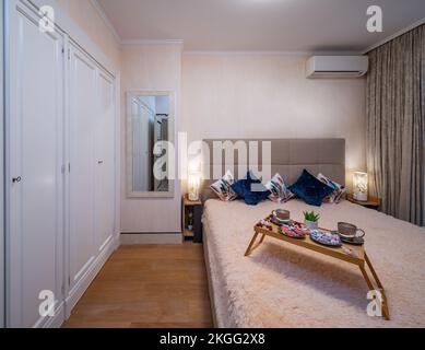 Moderne Innenausstattung des Schlafzimmers. Tablett mit Frühstück auf dem Bett. Weiße Garderobe Stockfoto