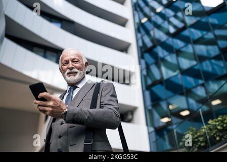 Low-Angle-Porträt eines leitenden Geschäftsführers mit Smartphone. Ältere Männer tragen einen Anzug, der während einer Geschäftsreise eine Laptop-Tasche mit sich führt Stockfoto