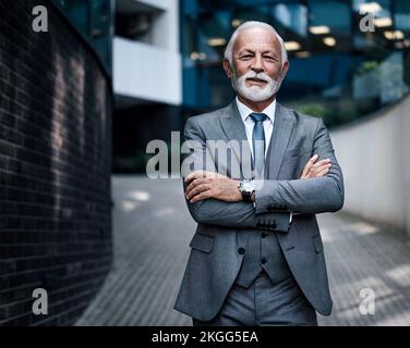 Porträt eines selbstbewussten älteren Unternehmertums mit verschränkten Armen. Ein lächelnder Geschäftsmann steht auf einem Fußweg vor einem modernen Bürogebäude Stockfoto