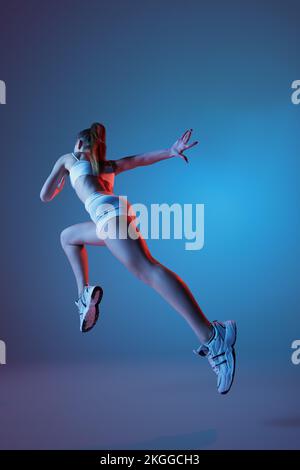 Rückansicht eines jungen Sportlers in weißer Sportbekleidung, das isoliert über einen abgestuften blauen violetten Hintergrund in Neonlicht wegläuft. Fitness, Hobby, gesund Stockfoto