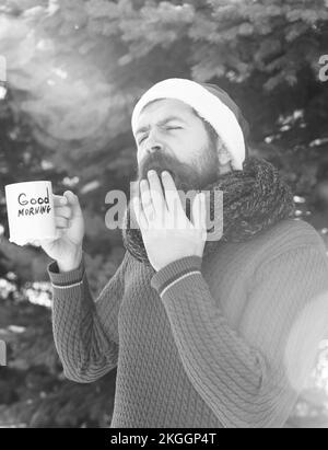 Lustig schöner Mann in weihnachtsmann Hut, bärtige Hipster mit Bart und Schnurrbart, leidet unter heißem Getränk in Tasse mit guten Morgen Text auf sonnigen Winter Stockfoto