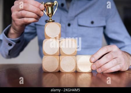 Geschäftsmann, der große Holzblöcke mit einer Siegertrophäe arrangiert. Geschäftserfolg Stockfoto