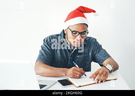 Laptop, weihnachtsmütze und Geschäftsmann schreiben Notizen, während er für ein Projekt in einem Studio recherchiert. Schreibtischmitarbeiter, Techniker und kreativer männlicher Angestellter Stockfoto