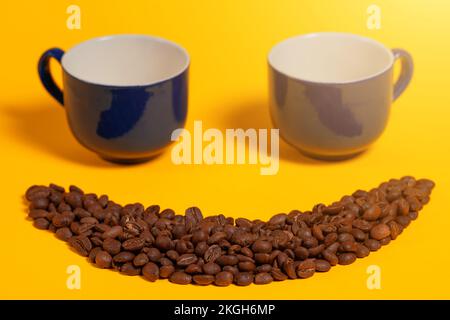 Fröhliches Emoticon aus Kaffeebohnen auf gelbem Hintergrund, Smiley aus Kaffee und zwei leere Tassen als Augen. Stockfoto